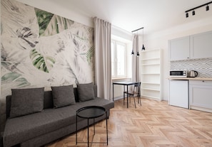 Ein modernes Wohnzimmer mit einer Couch, einem kleinen runden Tisch, einem Esstisch mit Stühlen und einer Küchenzeile mit Schränken und Geräten.