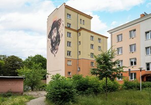 Ein mehrgeschossiges Gebäude mit Wandgemälde, umgeben von Grünflächen und an ein weiteres Gebäude anliegend.