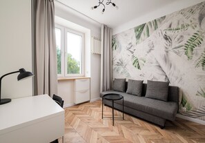 A room with a herringbone floor, a grey sofa, a small round table, a desk with a lamp, and a window with curtains. - Żytnia City Comfort Studio #5B (Warszawa)