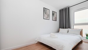 A modern bedroom with a wooden floor, a bed with white bedding, a window with curtains, and two framed pictures on the wall. - Łódź Downtown Charm Apartment (Łódź)