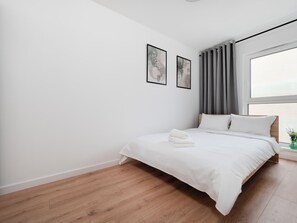 Ein modernes Schlafzimmer mit Holzboden, einem Bett mit weißer Bettwäsche, einem Fenster mit Vorhängen und zwei gerahmten Bildern an der Wand.