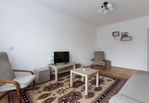 Ein Wohnzimmer mit einer Couch, einem Fernseher auf einem Schrank, einem kleinen Tisch, einem Sessel und einem gemusterten Teppich.