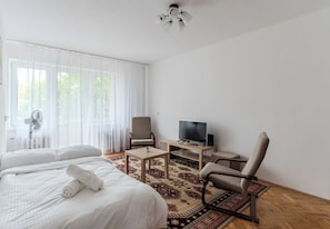 Ein Hotelzimmer mit einem Bett, zwei Stühlen, einem Fernseher, einem kleinen Tisch und einem großen Fenster mit durchsichtigen Vorhängen.