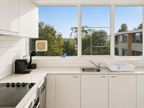 Eine moderne Küche mit weißen Schränken, einem eingebauten Backofen, einer Kaffeemaschine und einem Fenster mit Blick auf Bäume und Gebäude.