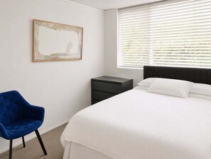 Ein Schlafzimmer mit einem Bett, einem Sessel, einem Nachttisch, einem Fenster mit Jalousien und einem gerahmten Bild an der Wand.