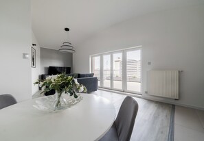 A modern dining area with a white table, grey chairs, a vase with flowers, a black TV, and a large window with a view of buildings. - Błażeja Modern Apartment (Poznań)
