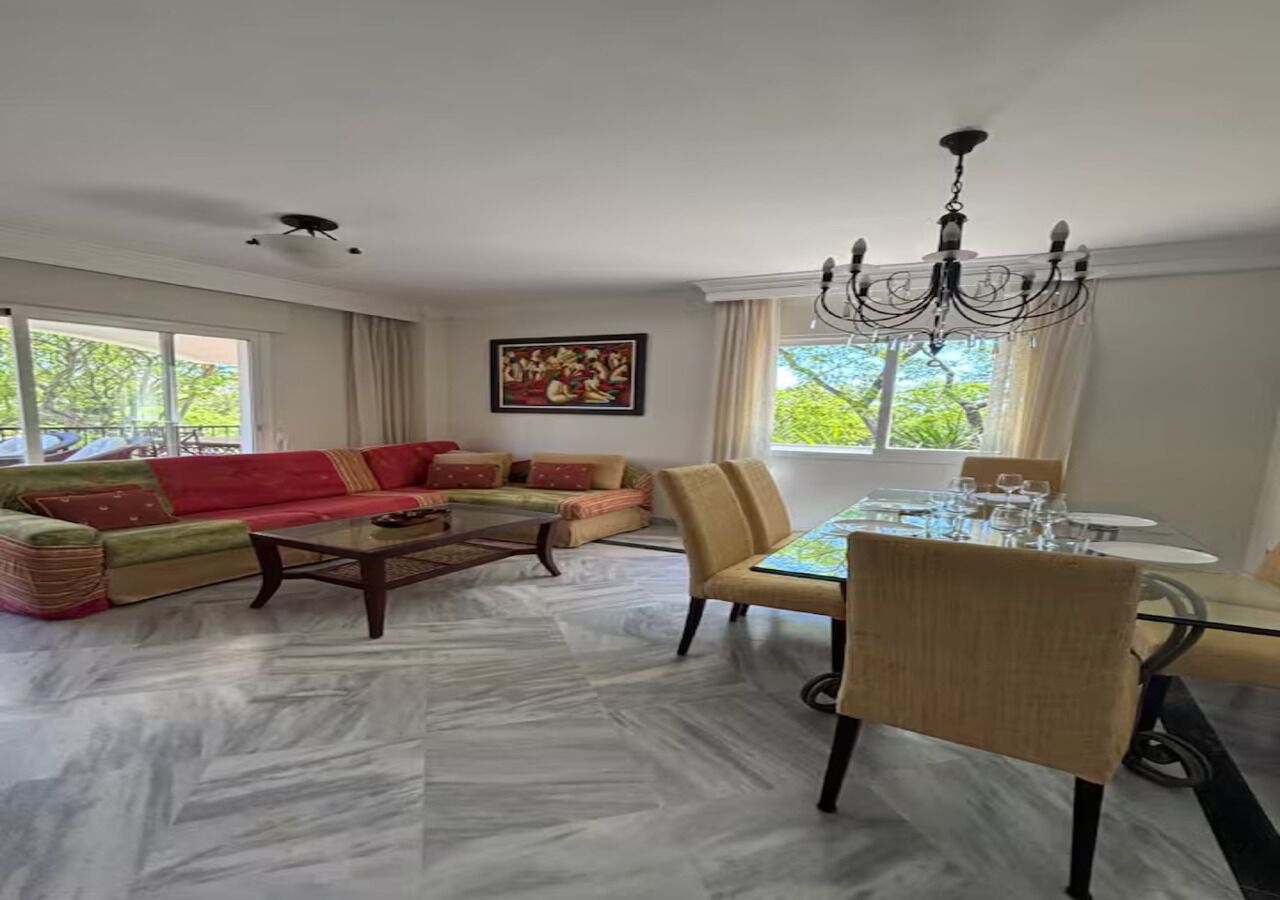 Ein Wohnzimmer mit einer Sofagarnitur, einem Esstisch mit Stühlen, einer Chandelüster und einem Gemälde an der Wand.