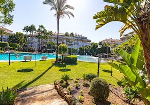 A pool area with a clear sky, palm trees, and a building in the background. - Relax in Banus - 3 bedrooms, pools, paddle - Pass the Keys (Marbella)
