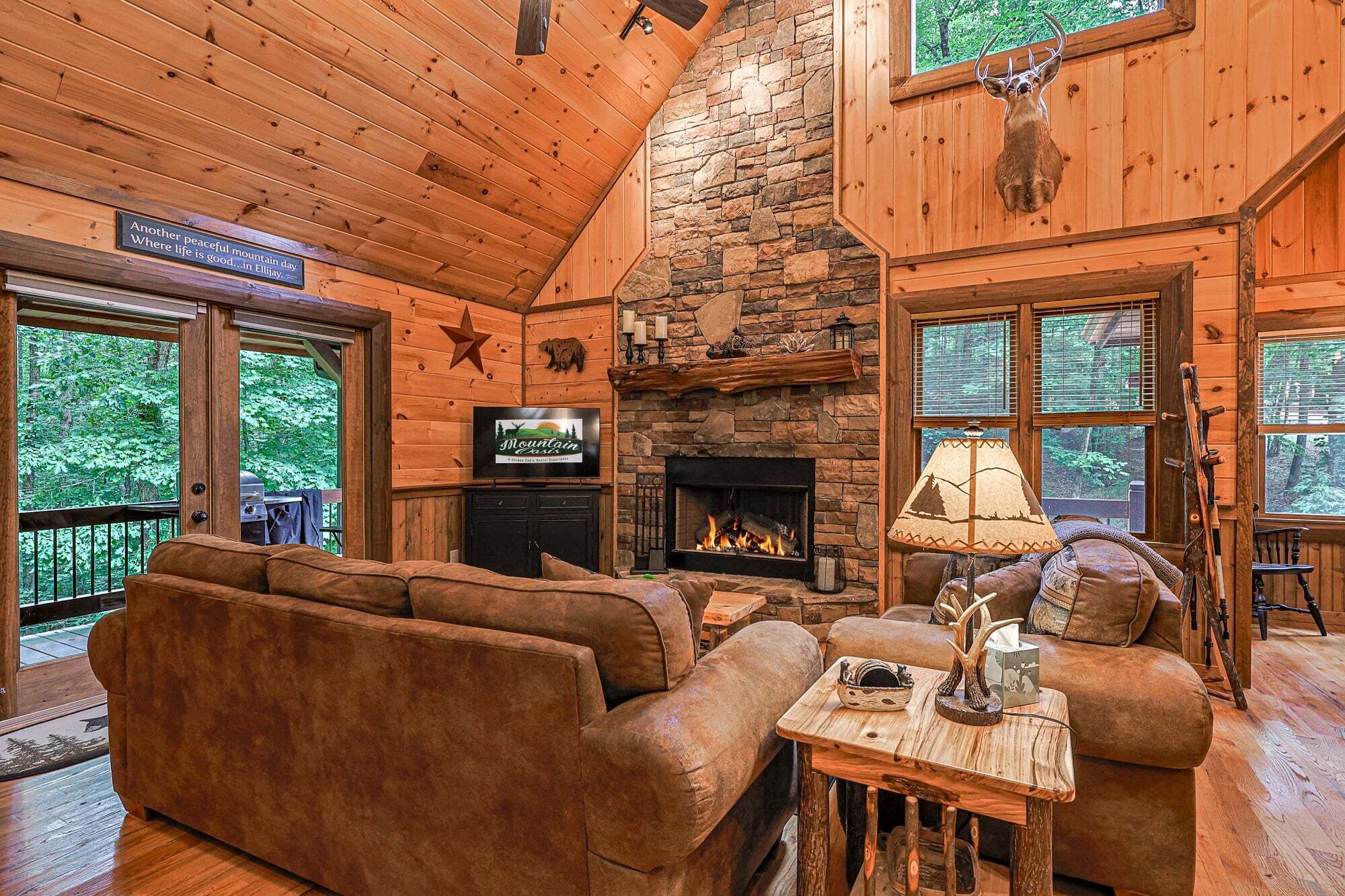 A cozy cabin interior with a stone fireplace, wooden walls, a television, and a fireplace mantel with decorative items.