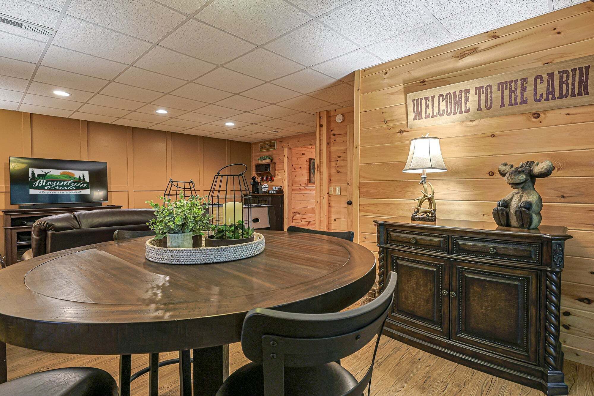 A cabin interior with a wooden wall, a round dining table, chairs, a television, and a cabinet with a lamp.