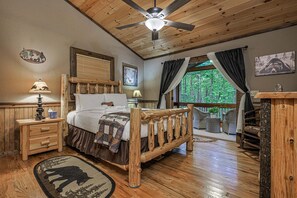 Ein rustikales Schlafzimmer mit einem hölzernen Bett, einem Deckenventilator, einem Fenster mit Blick auf Bäume und einem Nachttisch aus Holz mit einer Lampe.