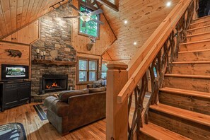 Ein Interieur einer Holzhütte mit Kamin aus Stein, einer Holztreppe und einem Ledersofa.