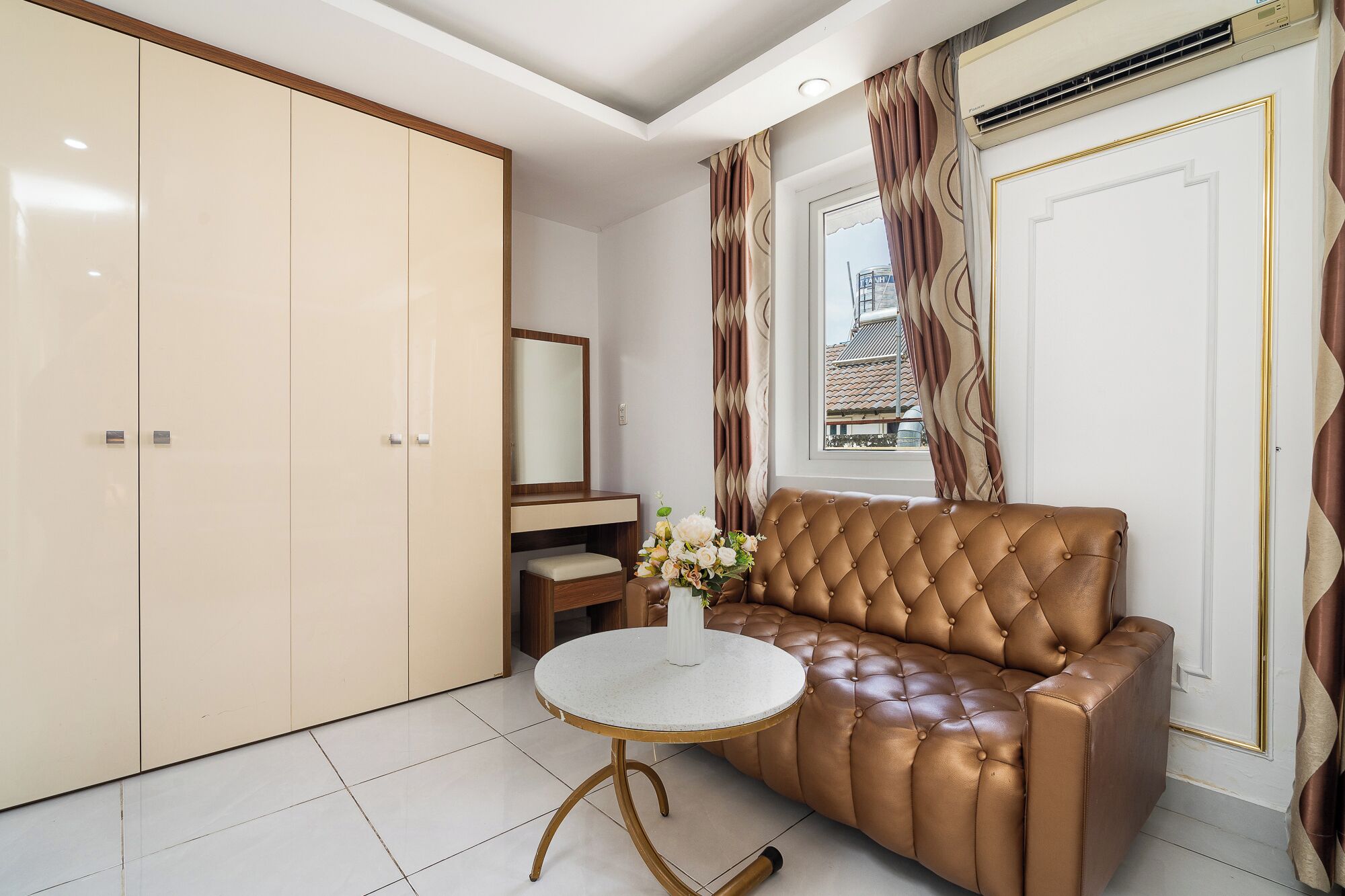 A room with a brown leather sofa, a round table with a white top, a mirror, a wardrobe, and a window with curtains.