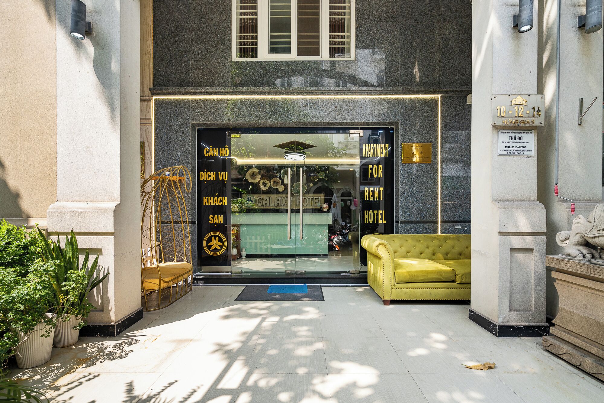 Entrada de hotel con puerta de cristal, mostrador de recepción, un sofá amarillo y una estatua.