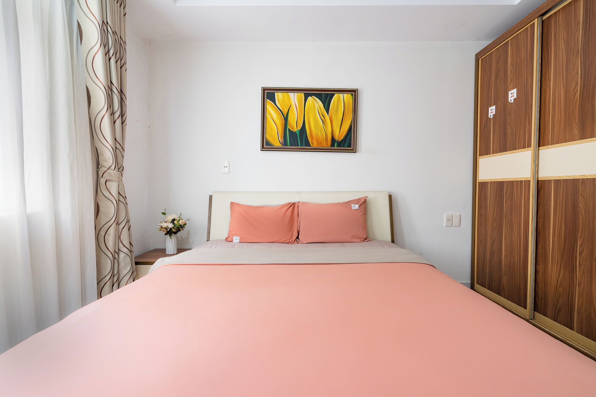 A bedroom with a bed, a painting, a wooden wardrobe, and a nightstand with a vase of flowers.