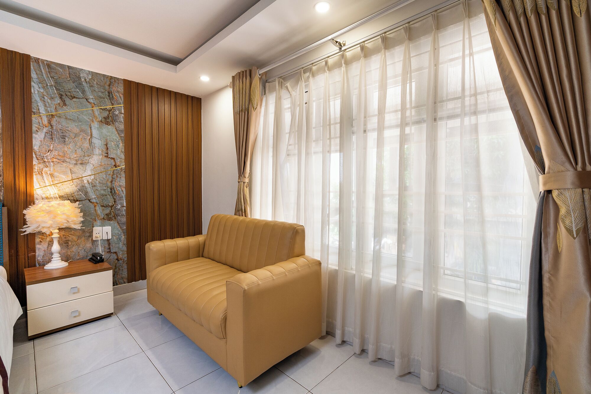 Una sala de estar moderna con un sofá, una lámpara, una pared con un diseño tipo mármol y cortinas translúcidas.
