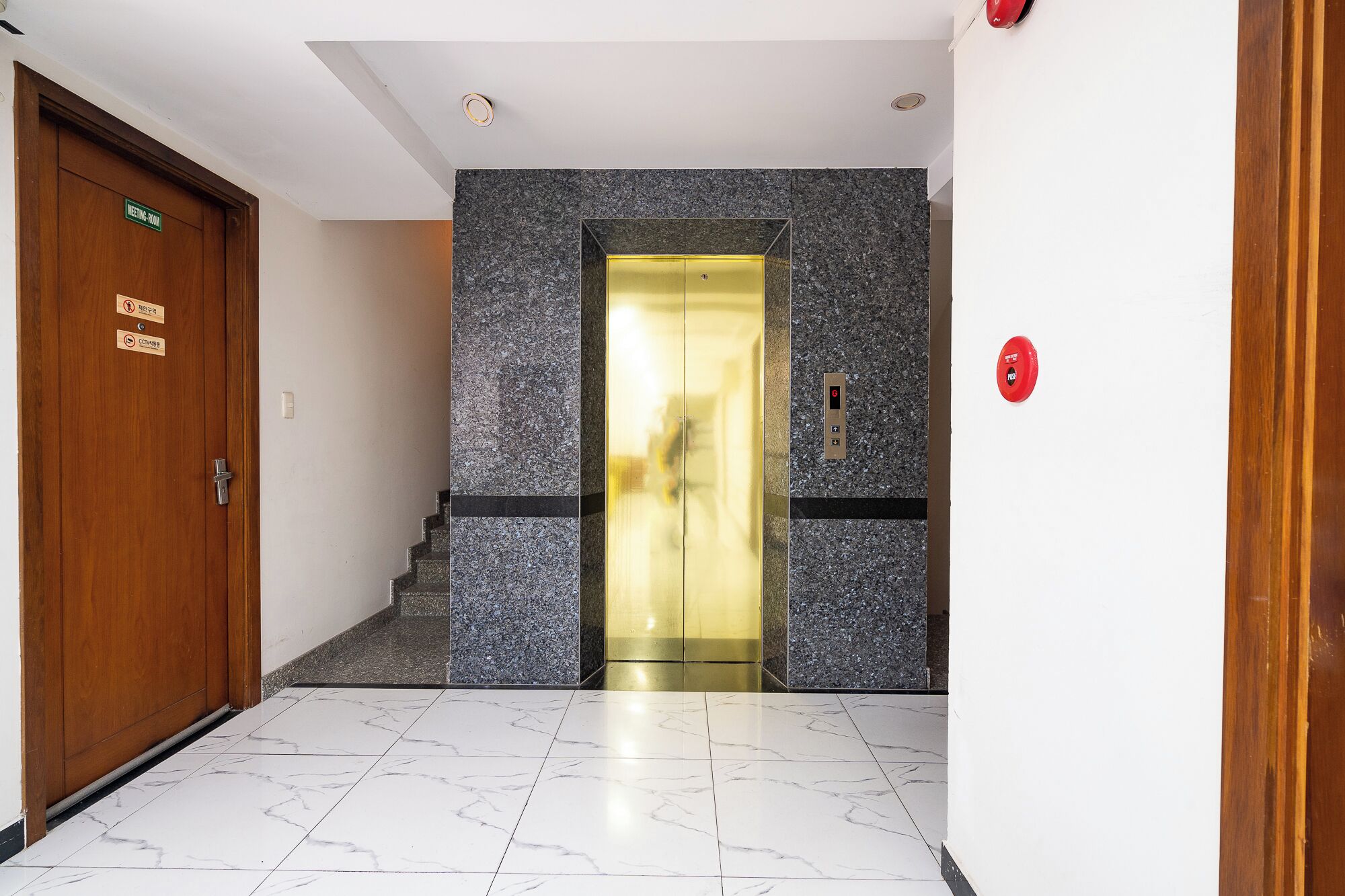 Un vestíbulo de hotel con piso de mármol, una puerta de madera con el letrero 'Sala de Reuniones' y un moderno ascensor con panel de control.
