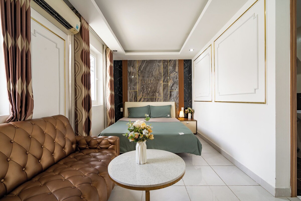 A bedroom with a tufted sofa, a round table, a bed with green bedding, and a marble wall.