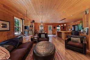A cabin interior with a wooden ceiling, a sofa, a leather ottoman, a dining area, and a kitchen.