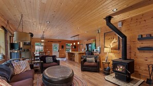 A cozy cabin interior with a wooden ceiling, a fireplace, a sofa, a dining area, and a kitchen. - Lake Pend Oreille Escape HotTub, GameRoom & Views (Sagle)