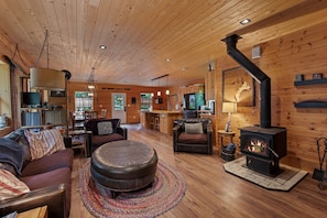 A cozy cabin interior with a wooden ceiling, a fireplace, a sofa, a dining area, and a kitchen.