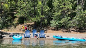 A lakeside setting with three blue Adirondack chairs, a blue inflatable raft, and a small boat with 'NAUTICAL' written on it. - Lake Pend Oreille Escape HotTub, GameRoom & Views (Sagle)