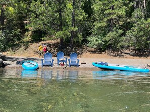 A lakeside setting with three blue Adirondack chairs, a blue inflatable raft, and a small boat with 'NAUTICAL' written on it.