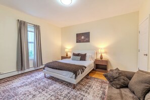 Quarto com cama, luminárias de cabeceira, janela com cortinas, tapete e um quadro na parede.