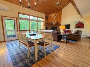 Un vaste espace de vie doté d’un plancher en bois, d’une table à manger avec des chaises, d’une cheminée, d’un téléviseur et d’une grande fenêtre offrant une vue sur la verdure.