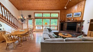 Een gezellige woonkamer met een open haard, houten vloeren, een eethoek met een houten tafel en stoelen, en grote ramen met uitzicht op groen.