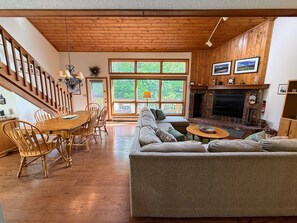Ein gemütliches Wohnzimmer mit Kamin, Holzböden, einer Essecke mit Holztisch und -stühlen sowie großen Fenstern mit Blick auf Grünflächen.