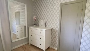 Una habitación con piso de madera, un aparador blanco con cajones, un espejo, una puerta y papel tapiz estampado.
