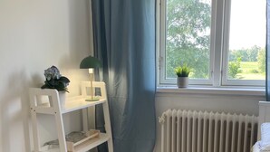 Una habitación con un escritorio blanco, un radiador, un ventanal con vista a follaje, y una alfombra gris.