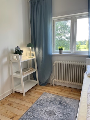 A room with a white desk, a radiator, a window with a view of greenery, and a grey rug.