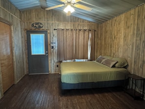 Quarto com paredes de madeira, cama com roupa de cama verde, ventoinha no teto e porta com janela.