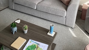 A grey sofa with patterned cushions, a framed abstract painting, a radiator, and a small table with a notebook, a map, and a can.