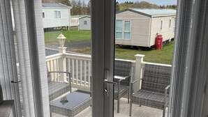 A room with a sliding glass door leading to an outdoor deck with a table and chairs, a view of a mobile home, and a grassy area.