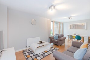 A modern living room with a sofa, a coffee table, a TV, and a dining area with chairs. - 1-bedroom Apartment Central Milton Keynes Emerald House (Moffat)