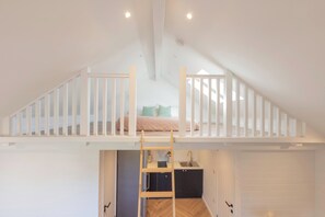 Dormitorio tipo loft con cama litera, una escalera de madera, zona de cocina y baño.