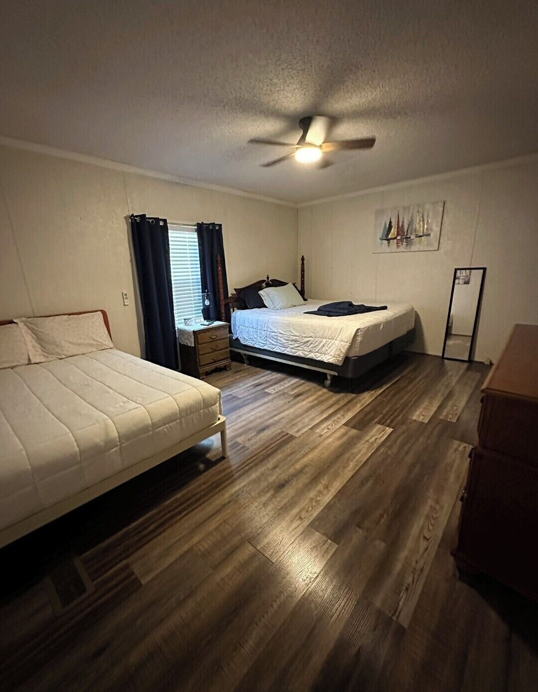 A room with two beds, a ceiling fan, a window with curtains, a small table, and a mirror.