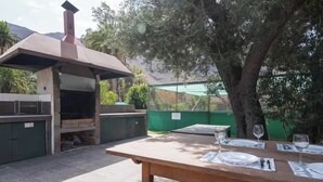 Zona de barbacoa al aire libre con una parrilla, una mesa lista para comer y una valla verde al fondo.