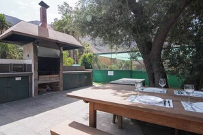 Ein Grillbereich im Freien mit Grill, Esstisch und grünem Zaun im Hintergrund.