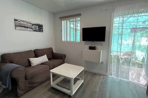 Ein Wohnzimmer mit braunem Sofa, einem weißen Couchtisch, einem flachbildfernseher an der Wand und einem Fenster mit Blick ins Freie.