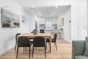 A modern kitchen with white cabinets, a dining area with a wooden table and chairs, and a living room with a green sofa. - The Casey - Effortless Luxury (Orange)