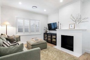 A living room with a sofa, ottoman, fireplace, TV, and a bookshelf. - The Casey - Effortless Luxury (Orange)
