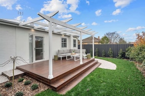 A backyard with a wooden deck, a white pergola, a gray sofa, and a small table. - The Casey - Effortless Luxury (Orange)