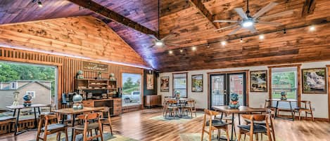 A spacious dining area with wooden tables and chairs, a bar area with shelves, and large windows offering views of the outdoors.
