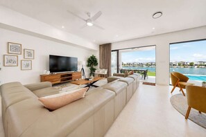 Ein modernes Wohnzimmer mit einer großen Couch, einem Flachbildfernseher und Blick auf ein Gewässer durch bodentiefe Fenster.