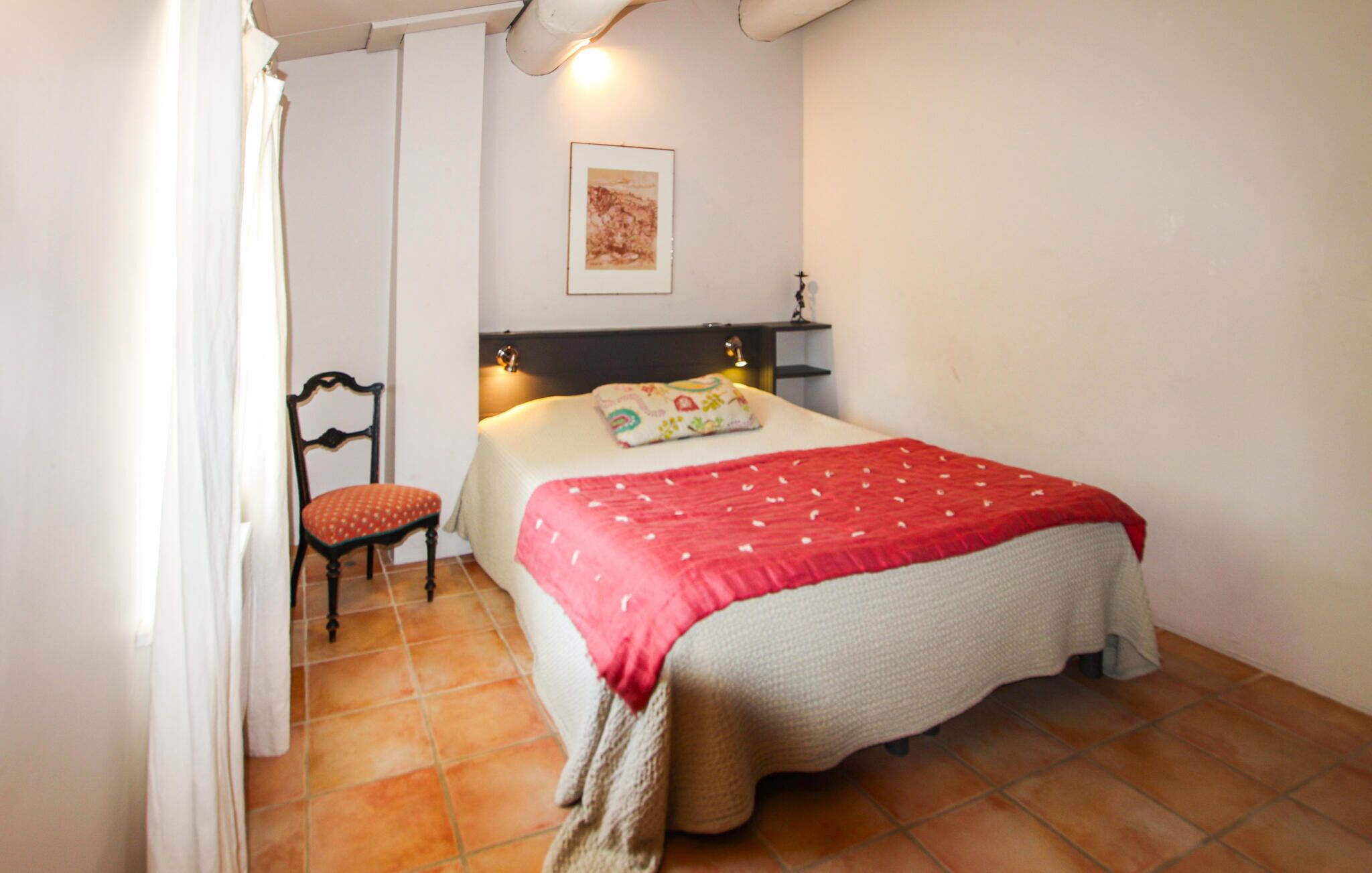 A bedroom with a bed, a chair, a nightstand, a wall-mounted lamp, and a framed picture.