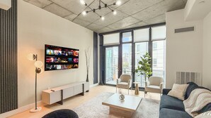 A modern living room with a flat-screen TV, a coffee table, sofas, and a large window with a city view. - Apartmint at N 2nd St Philadelphia (Philadelphia)